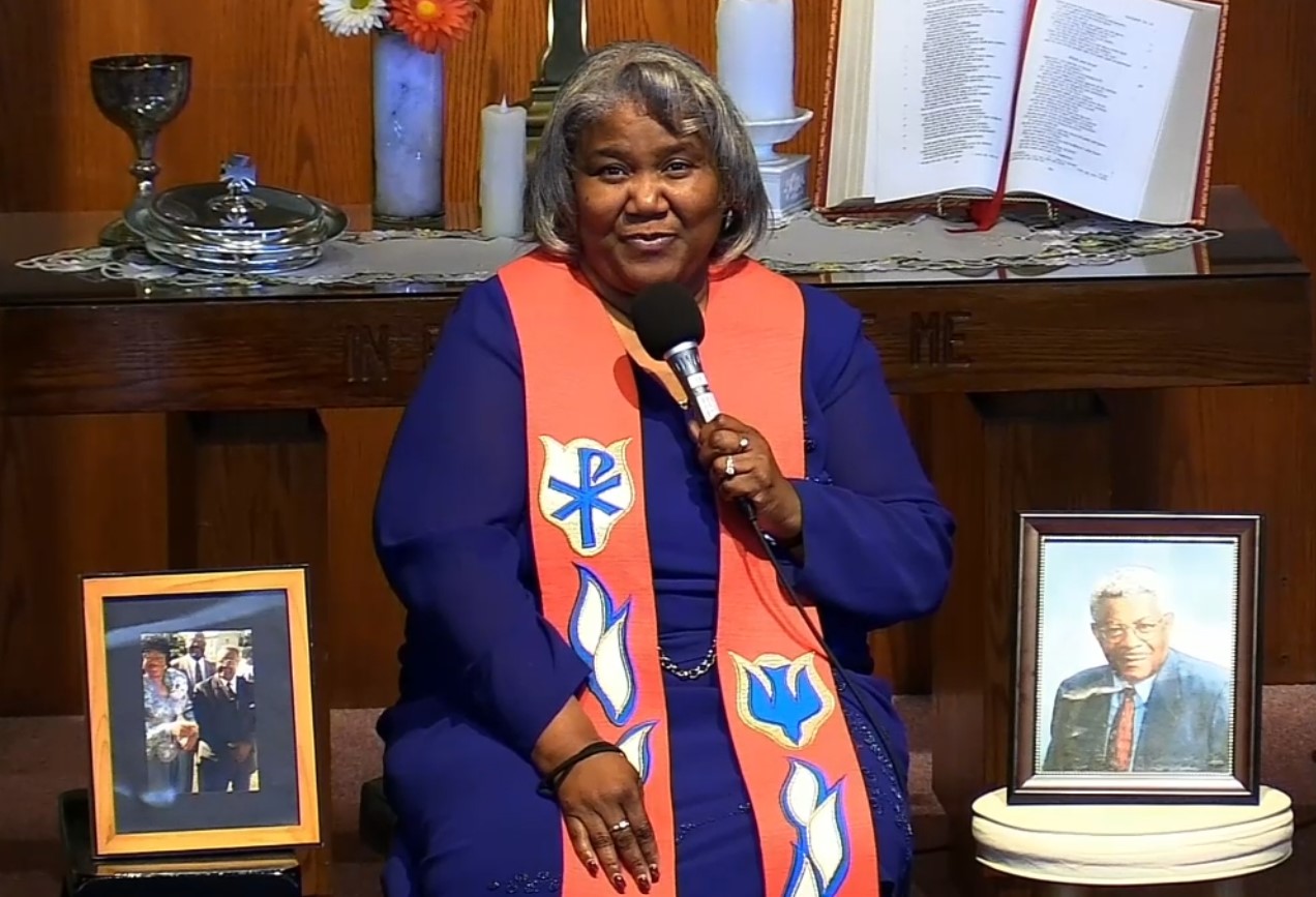 Speaker wearing a royal blue long sleeve dress, and a coral Christian prayer shawl, with two important representative photos on either side of her, as well as candles on a mantle behind her and a bible.