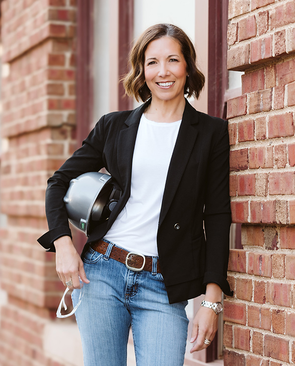 Barb-Allen headshot,brunette curled bob, grey hardtop hat, clear work glasses, white shirt, black suit jacket, blue jeans with brown belt, brick background.